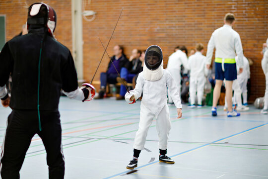 Little Kid Boy Fencing On A Fence Competition. Child In White Fencer Uniform With Mask And Sabre. Active Kid Training With Teacher And Children. Healthy Sports And Leisure.