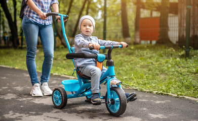 Young mother is pushing a child's tricycle with a toddler boy on a walk. Concept of learning to ride a bike and having fun with your family - obrazy, fototapety, plakaty