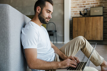Image of young thinking man working with laptop while sitting on floor