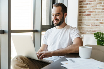 Image of handsome joyful man smiling and working with laptop