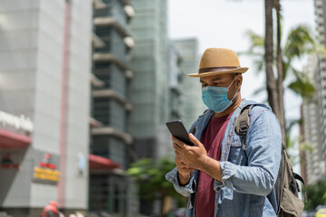 Young asian backpacker traveling in urban city using smartphone to search the location to travel. He wearing face mask for protect virus safety travel.