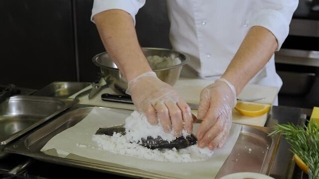 Close-up of the chef sprinkling salt on raw fish before baking in the oven. The process of cooking Dorado fish in a large amount of salt in a professional kitchen.