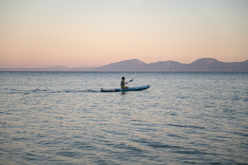 Fototapeta premium Beautiful view of young boy sitting on sup board paddling on calm evening sea