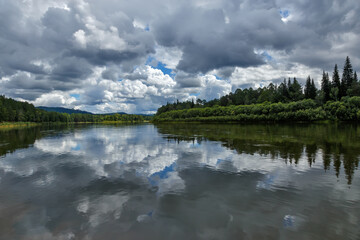 A natural landscape with clouds reflecting on the surface of a calm river with green forest on its banks in Siberia