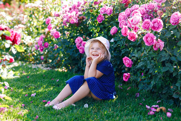 Portrait of little toddler girl in blossoming rose garden. Cute beautiful lovely child having fun with roses and flowers in a park on summer sunny day. Happy smiling baby.