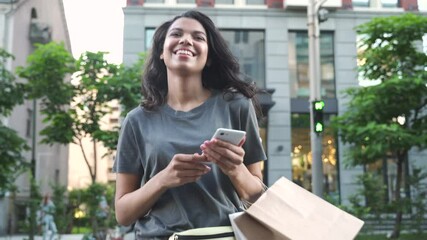Smiling happy young adult African American ethnic girl shopper customer walking on city street holding shopping bags and smart cell phone using mobile fashion sale ecommerce app near shop mall store.