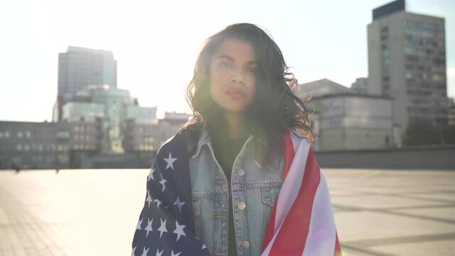 Proud serious young African American woman standing on street wrapped in usa flag look at camera outdoor. Black people for democracy, fair elections against racism for rights equality in United States