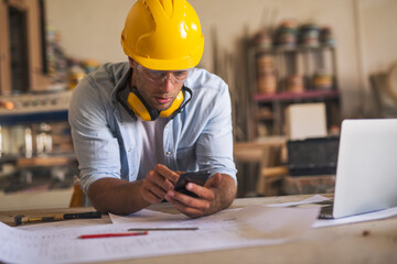 Close up of young carpenter leaned on his work desk using mobile phone