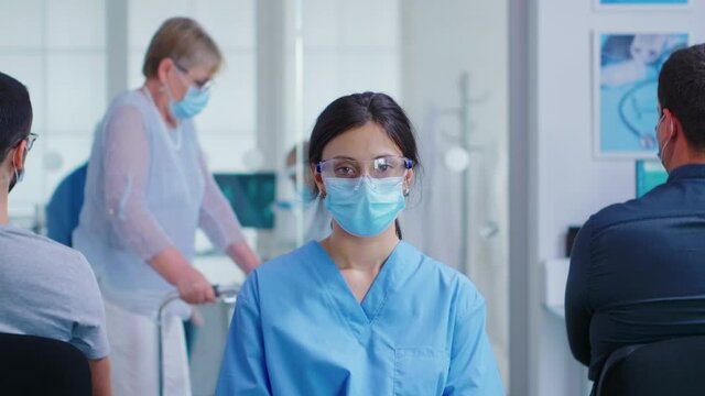 Female nurse with face mask against coronavirus in hospital waiting area looking at camera. Senior woman with walking disability using zimmer frame.