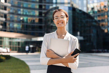 Portrait of a young business woman in glasses with a smile holding a laptop and smartphone in her hands, stands on the street, against the background of a business center