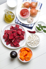 Raw beef, white beans, onions, carrots, garlic, bay leaf, pepper, tomato paste and olive oil on a white table