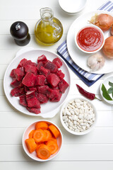 Raw beef, white beans, onions, carrots, garlic, bay leaf, pepper, tomato paste and olive oil on a white table