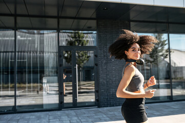 Fototapeta na wymiar Charming African-American young woman is running outdoors. A girl with an afro hairstyle looks back at camera while jogs on the street