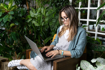 Concentrated female gardener in eyeglasses wear linen dress, sitting on chair in green house, working on laptop surrounded by exotic plants. Freelance