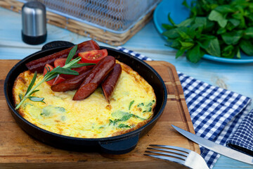 Omelet with sausage and tomatoes in a pan on the table. Appetizing breakfast. A sprig of rosemary, a wooden spice. Hunt sausages. Tasty food fried eggs.