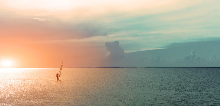 silhouette windsurfer sailing on windsurf board on sea at a beautiful sunset sky on background. Healthy active lfestyle, hobby, summer fun adventure.
