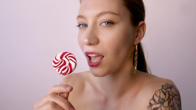 Beauty female tattooed model with red hair style and trendy beautiful makeup holding pink sweet colorful lollipop candy.Fashion woman having fun over background