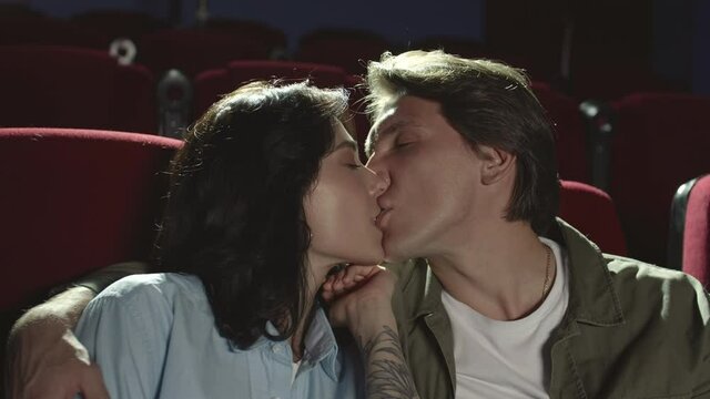 Medium shot of middle-aged Caucasian man kissing with tenderness his pretty girlfriend sitting nearby in cinema and watching film