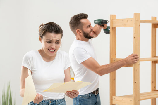 Frowning puzzled young woman looking at assembly plan with confusion while her annoyed husband assembling furniture