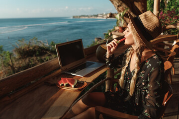 Pretty woman in straw hat works with a laptop on the beach cafe and eat watermelon