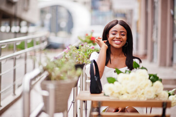 Beautiful african american girl holding bouquet of white roses flowers on dating in the city. Black businesswoman with bunch of flowers sitiing by the table in outdoor cafe.