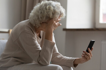 Sad elderly woman sit on sofa hold smartphone feels disappointed by received sms bad news, awful...