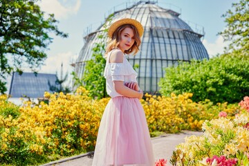Obraz na płótnie Canvas Gorgeous young woman in hat posing in a blooming garden. Beautiful girl dressed in pink skirt and white blouse. Sensual woman with perfect skin and with natural makeup. fashion and style concept.
