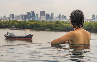 Fototapeta premium Side view of Short haired young woman relax in the roof top outdoor swimming pool and enjoying the city view Bangkok. Summer vacation concept.