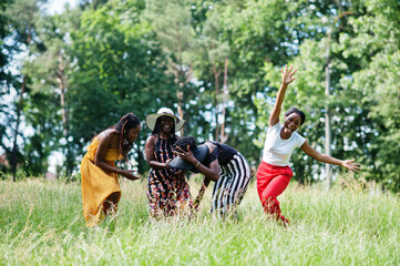 Group of four gorgeous african american womans wear summer hat jumping at green grass in park.