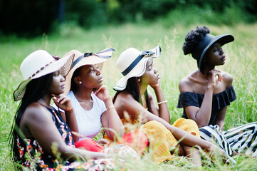 Group of four gorgeous african american womans wear summer hat sitting at green grass in park.