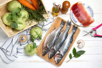 Raw sea bass, kohlrabi, carrots, thyme, garlic, bacon, bay leaf and olive oil on a white table