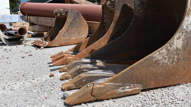 Baggerschaufeln auf Asphaltplatz, drei Schaufeln mit Baggerz&auml;hnen, metallene Gegenst&auml;nde als grafische Elemente	
