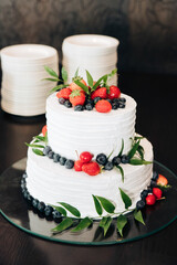 Festive wedding or other festive cake. Two-layer cake, white, decorated with fruit, strawberries, blueberries, eucalyptus leaves and other greens. Glass tray, dark background, behind the cake plates