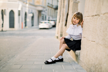 Outdoor fashion street city portrait of little Caucasian girl. Happy little girl sitting near the old building wall in ancient European city with blue door on the background