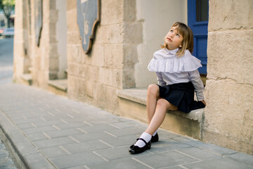 Outdoor fashion street city portrait of little Caucasian girl. Happy little girl sitting near the old building wall in ancient European city with blue door on the background