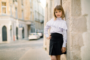 Front view of little blond girl in black and white clothes posing at the street city against the background of the wall of old building