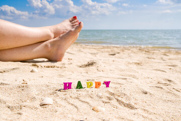 Selective focus on the word Happy from multicolored letters among sand and seashells. In the background there are beautiful female feet with a pedicure of bright red color and the sea. Vacation at sea