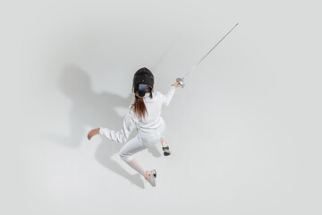 First. Teen girl in fencing costume with sword in hand on white background. Top view. Young female model practicing and training in motion, action. Copyspace. Sport, youth, healthy lifestyle.