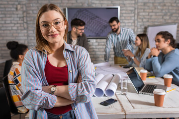 Portrait of confident young female leader standing and looking at camera while her colleagues working on architectural designs in background.