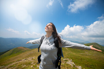A carefree happy female tourist with backpack is breathing deeply with open arms raised just reached a peak while hiking in the middle of hills surrounded by green nature.