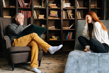 Happy red-haired young woman patient is telling about her mental health while lying in comfortable sofa during psychological consultation. Professional psychologist listening to patient at dark office