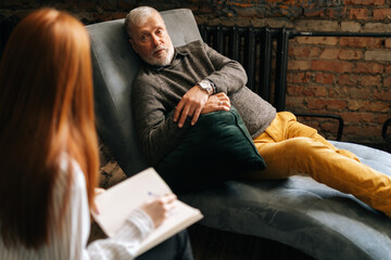 Young woman psychologist sitting at back to camera is talking to mature patient lying in comfy sofa in office making notes asking questions during individual counselling. Concept of mental care. 