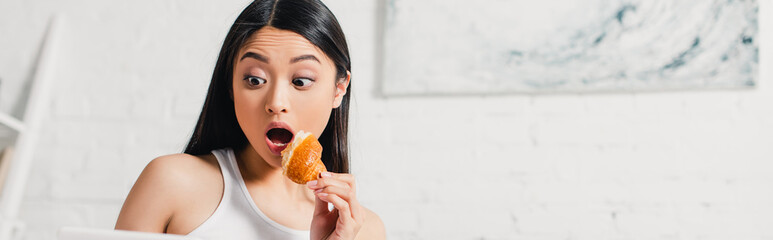 Panoramic shot of shocked asian woman holding piece of croissant at home