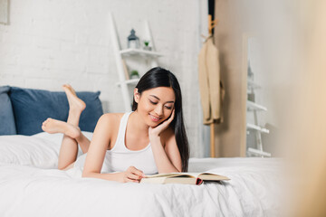 Selective focus of smiling asian girl reading book while lying on bed at home