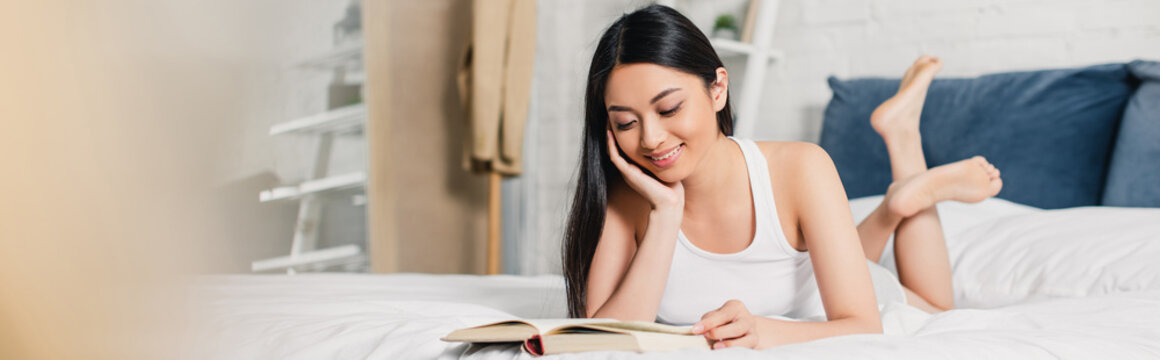 Horizontal image of smiling asian girl with hand near cheek reading book on bed