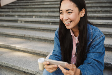 Image of asian woman playing game on cellphone while sitting at stair