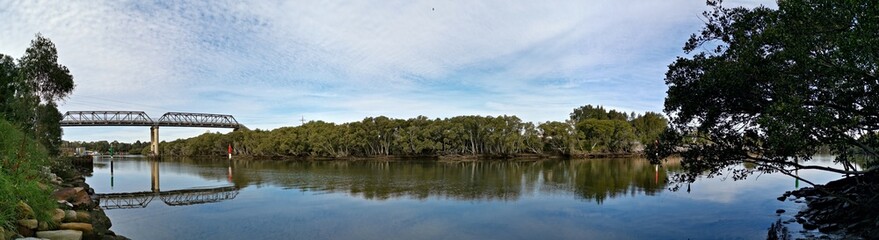 Beautiful panoramic view of a river with reflections of a tall pedestrian and water pipe  bridge, trees, deep blue sky and puffy clouds on water, Parramatta river, Rydalmere, Sydney, New South Wales, 