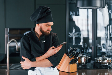 Professional young chef cook in uniform standing with phone on the kitchen