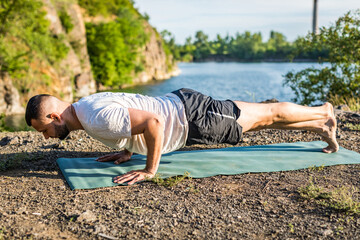 Fototapeta premium Morning and evening, a handsome young man doing yoga exercise on the shore of a quarry lake in nature in the summer outdoors. Healthy active lifestyle concept.