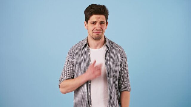 Young bunet man in casual outfit is pretending like crying and wiping his tears by hand while posing against blue studio background